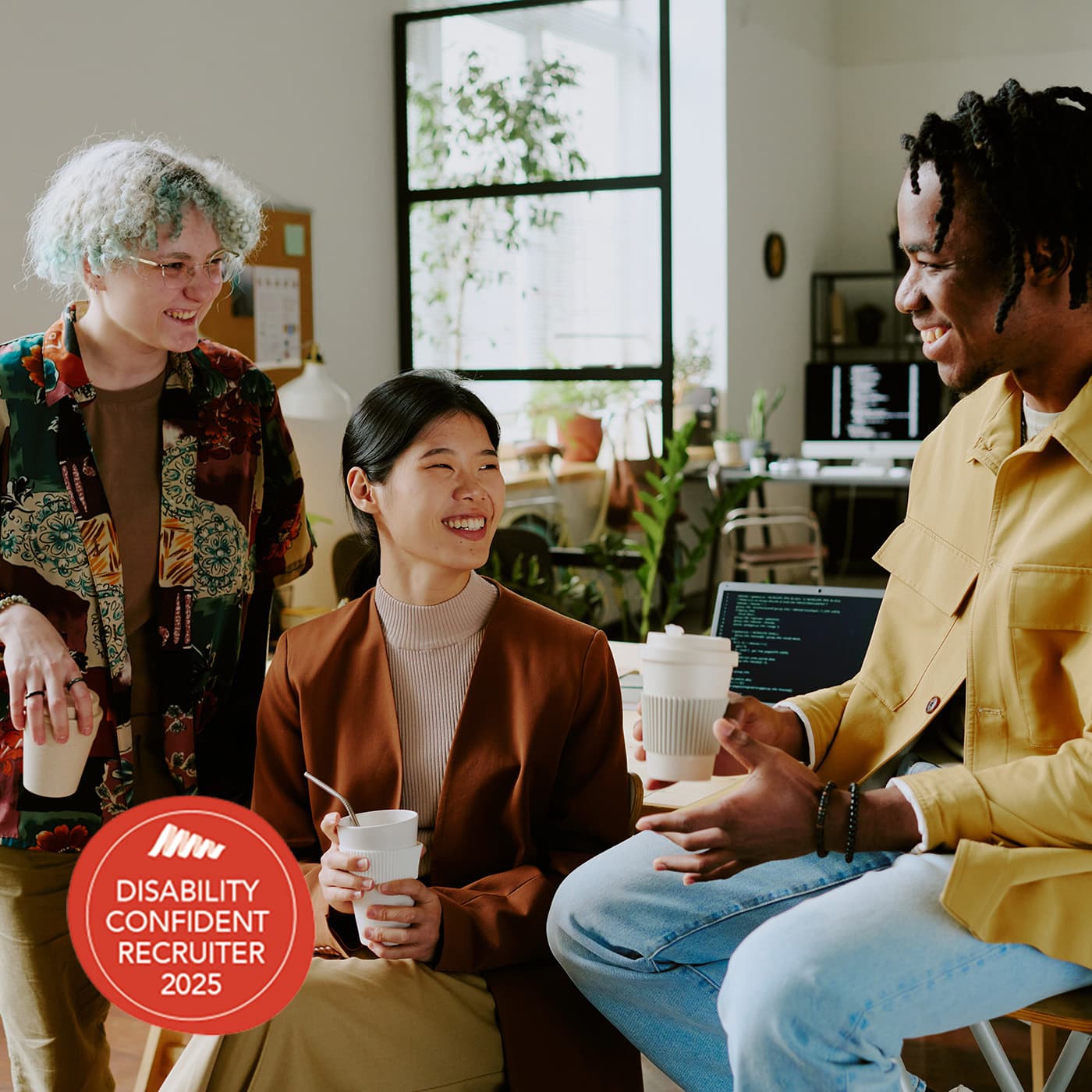 Three diverse male and female work mates wearing casual clothes enjoying coffee break at work in modern office. The Disability Confident Recruiter badge is overlaid at bottom left. Three diverse male and female work mates wearing casual clothes enjoying coffee break at work in modern office. The Disability Confident Recruiter badge is overlaid at bottom left.