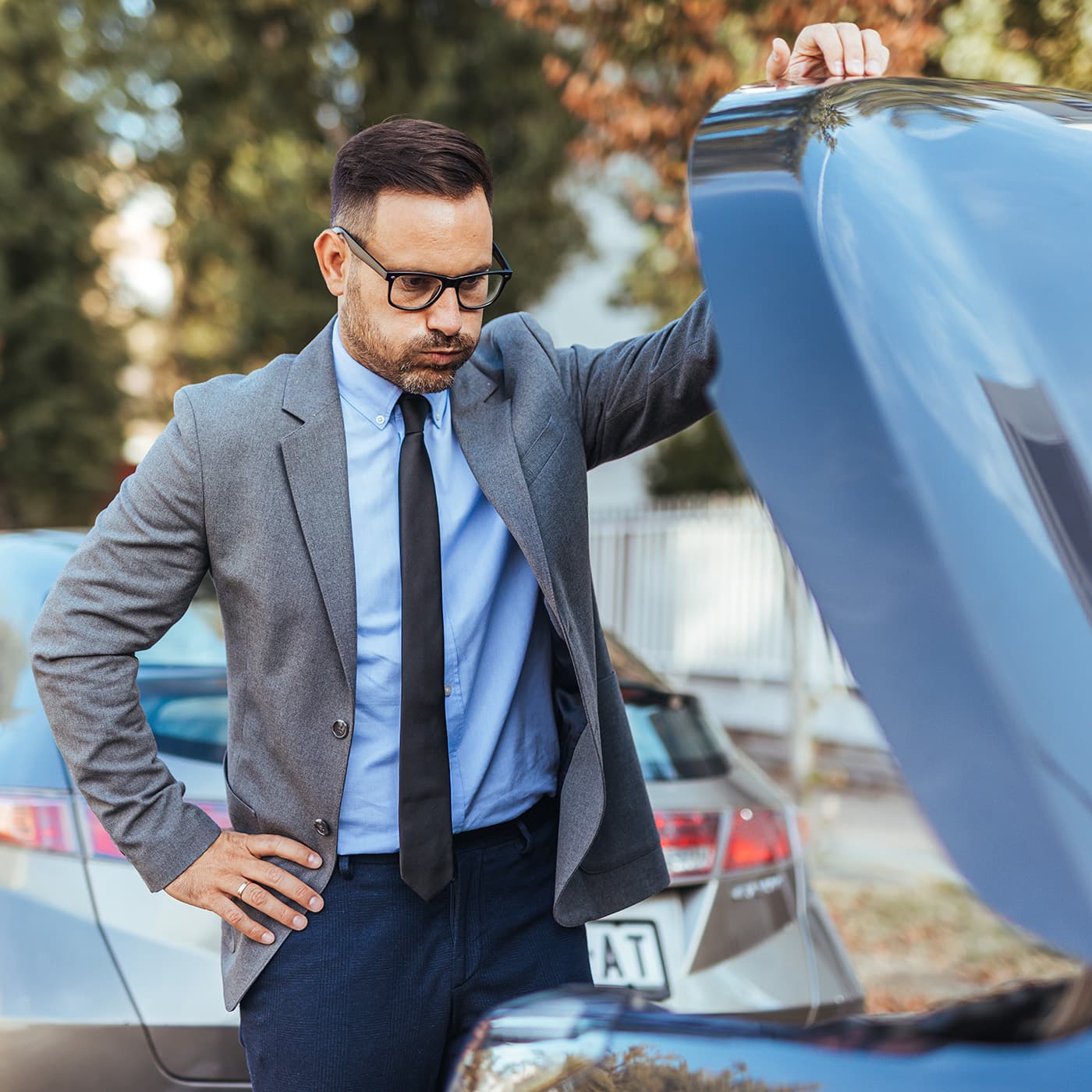 A concerned man in a suit inspects a car engine in a city setting, possibly dealing with a breakdown or mechanical issue. A concerned man in a suit inspects a car engine in a city setting, possibly dealing with a breakdown or mechanical issue.
