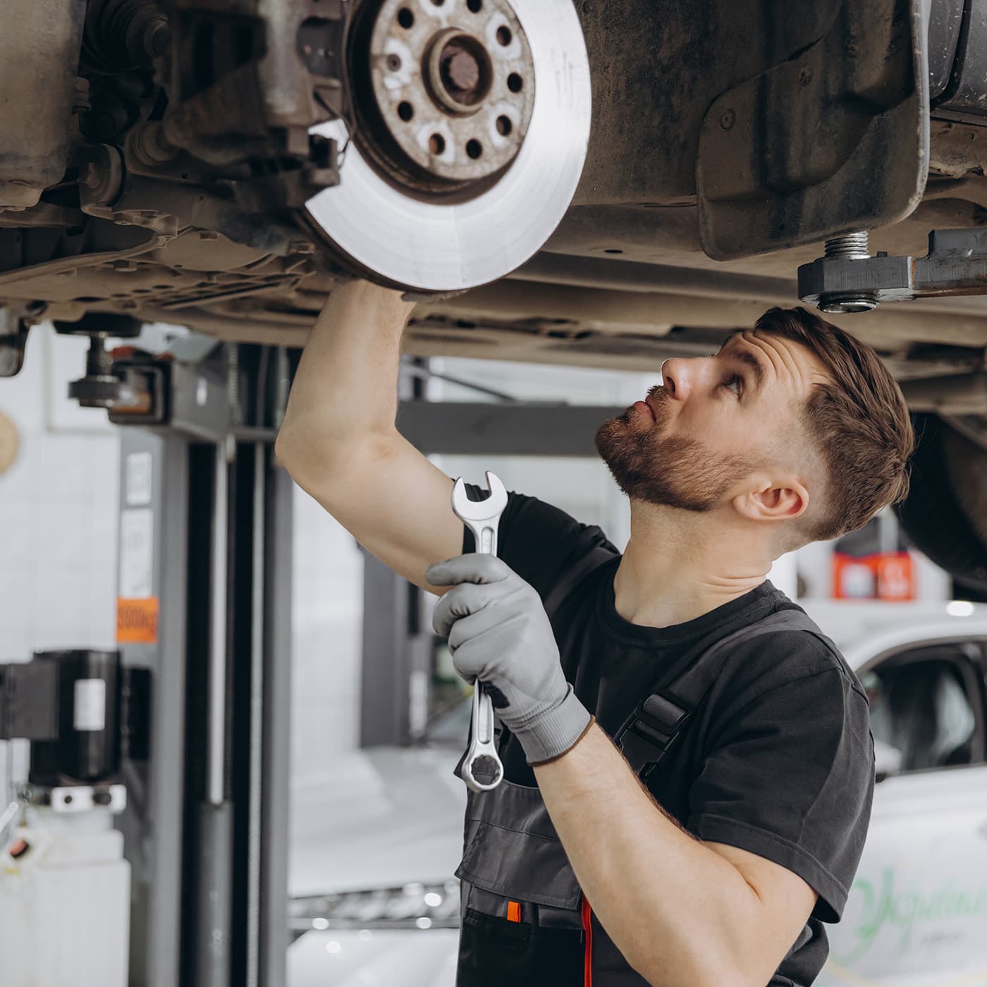 View from side of concentrated worker of car service checking running gear of automobile. Male mechanic in uniform and gloves standing under car and looking for breakage. View from side of concentrated worker of car service checking running gear of automobile. Male mechanic in uniform and gloves standing under car and looking for breakage.