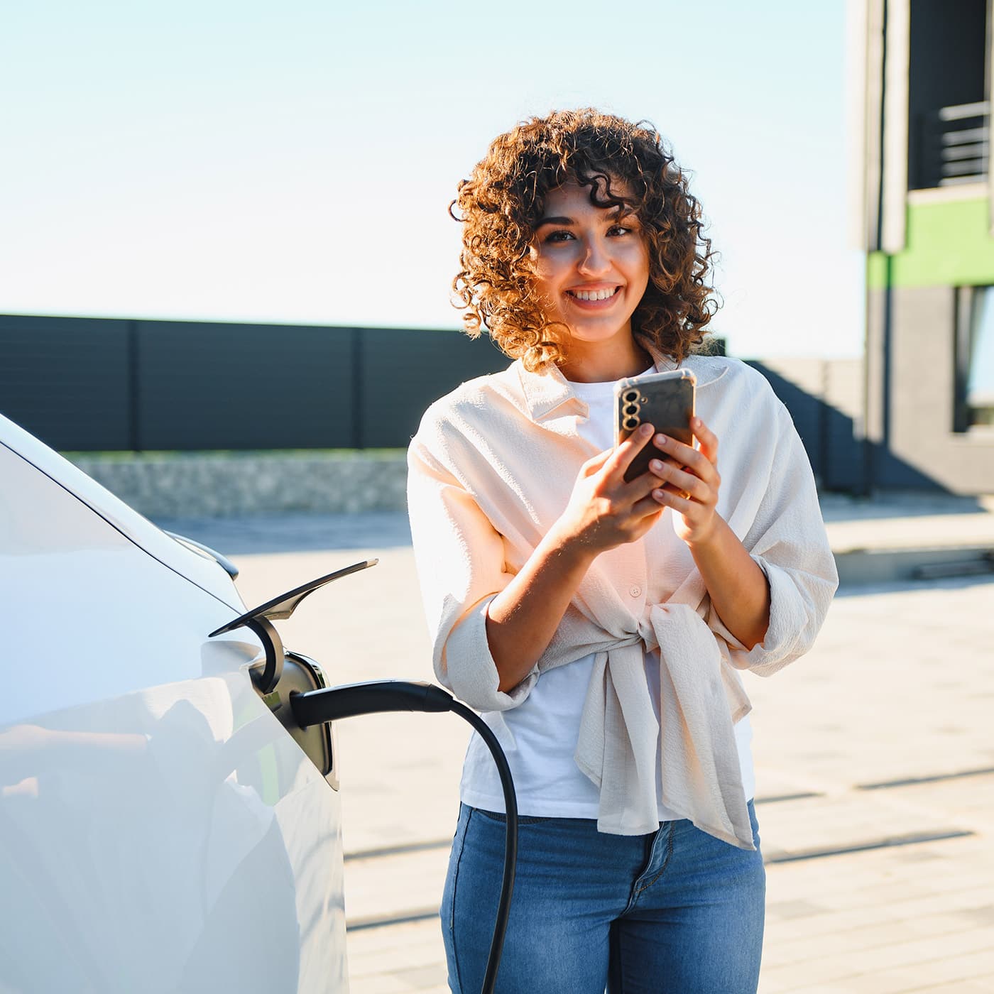 Smiling woman with curly brown hair using her smartphone while charging her EV in front of her house. Smiling woman with curly brown hair using her smartphone while charging her EV in front of her house.