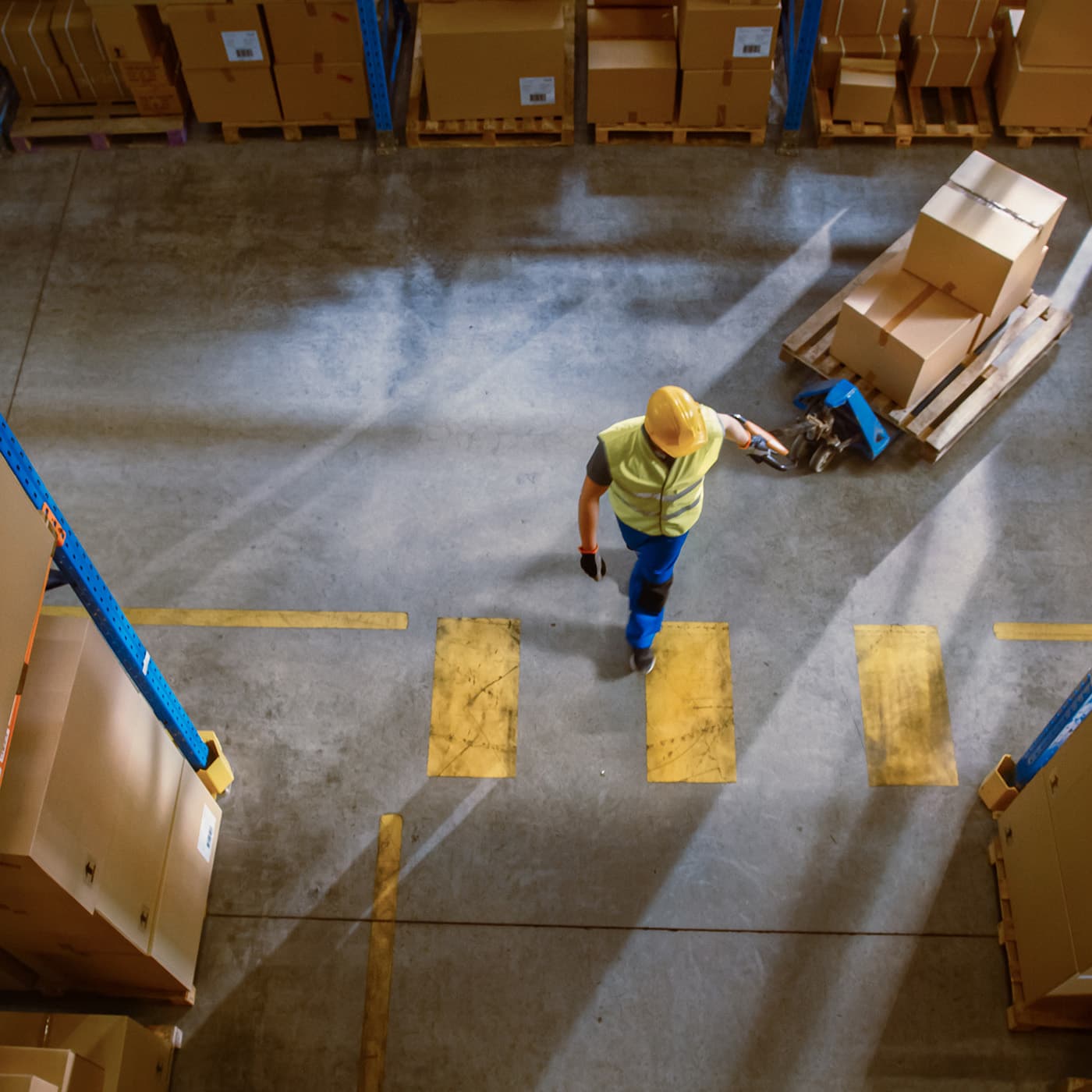 Aerial view: Worker moves boxes using manual pallet truck, walking between rows of shelves in retail warehouse. Aerial view: Worker moves boxes using manual pallet truck, walking between rows of shelves in retail warehouse.