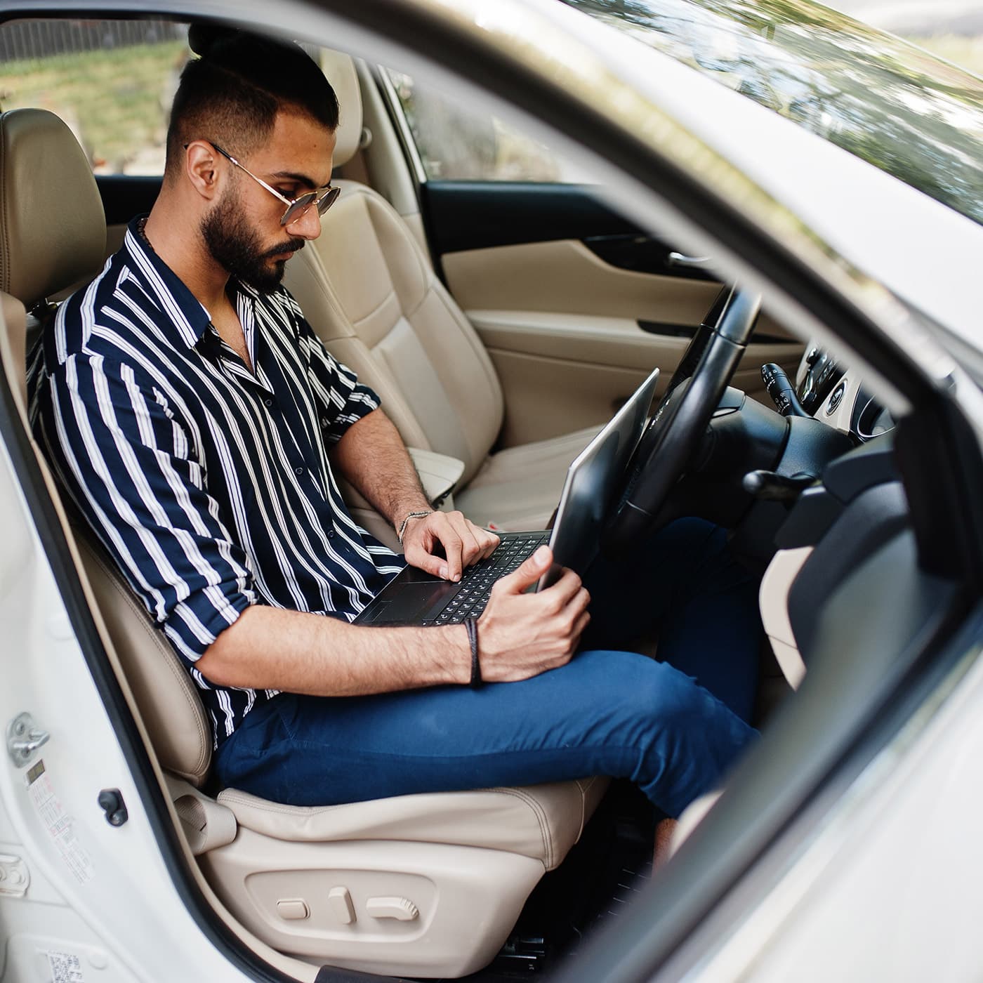A man in a striped shirt and sunglasses behind the wheel of his white vehicle with laptop. A man in a striped shirt and sunglasses behind the wheel of his white vehicle with laptop.