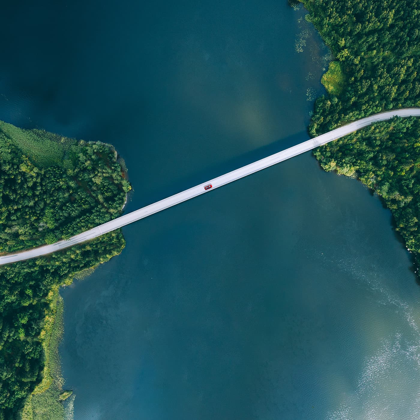 Aerial view of bridge road with red car over blue water lake or sea with island and green woods. Aerial view of bridge road with red car over blue water lake or sea with island and green woods.