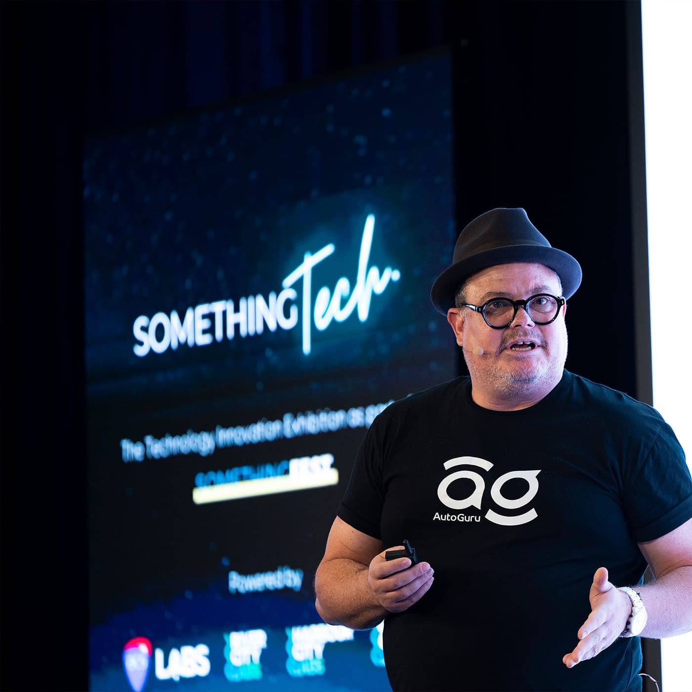 The image shows a photo of AutoGuru’s CEO, Eden Shirley, speaking at the Something Tech conference. Eden is wearing a black t-shirt with a white AutoGuru logo and a black hat. The image shows a photo of AutoGuru’s CEO, Eden Shirley, speaking at the Something Tech conference. Eden is wearing a black t-shirt with a white AutoGuru logo and a black hat.