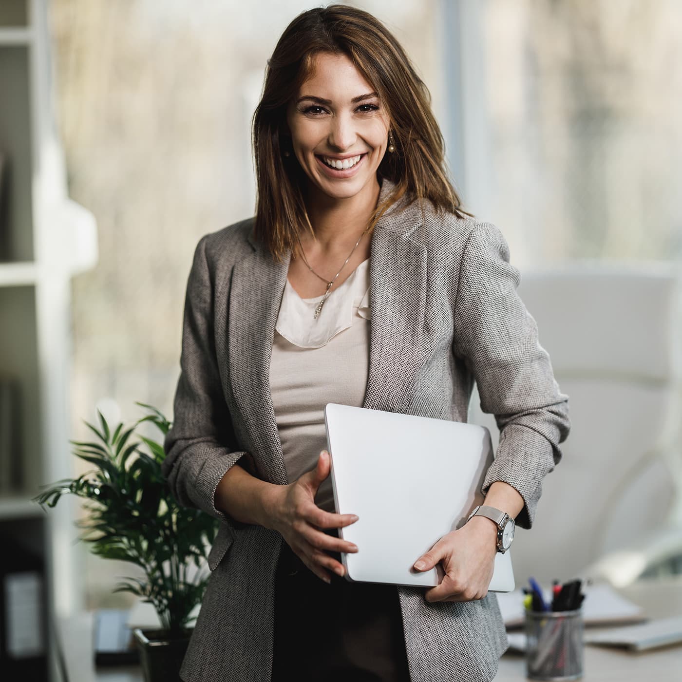 A young female Strategic Advisor holding laptop and standing in her office, smiling. A young female Strategic Advisor holding laptop and standing in her office, smiling.