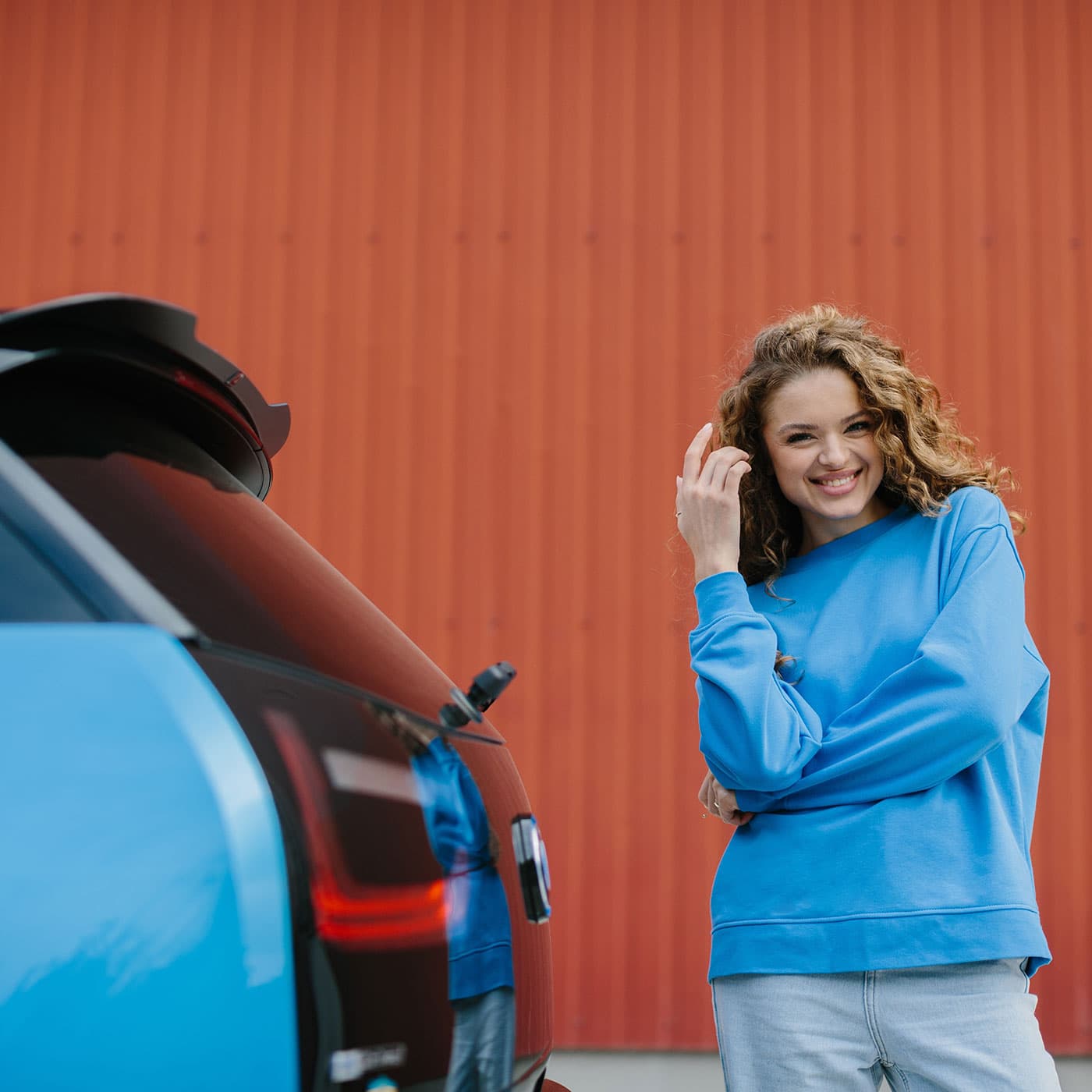 A smiling young woman wearing a blue sweatshirt and light coloured pants, standing at the back of her electric vehicle. Behind her is an orange wall. A smiling young woman wearing a blue sweatshirt and light coloured pants, standing at the back of her electric vehicle. Behind her is an orange wall.
