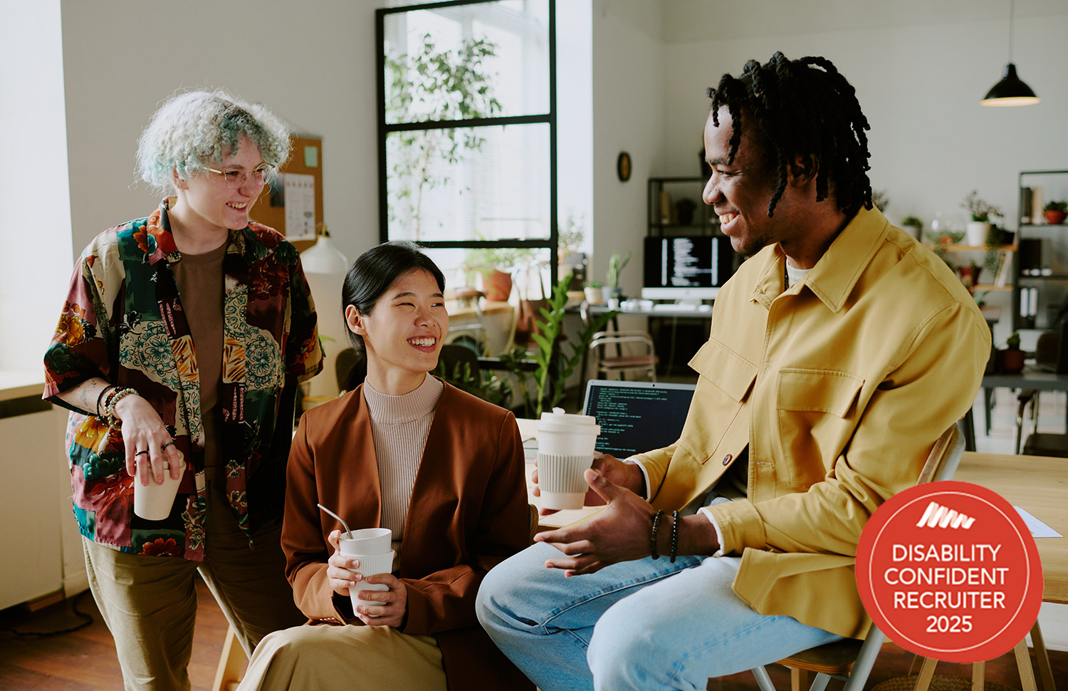 The image shows three people of different ages, genders and cultural backgrounds, smiling and sharing a coffee in the workplace. It also includes the Disability Confident Recruiter 2025 logo in the bottom right.
